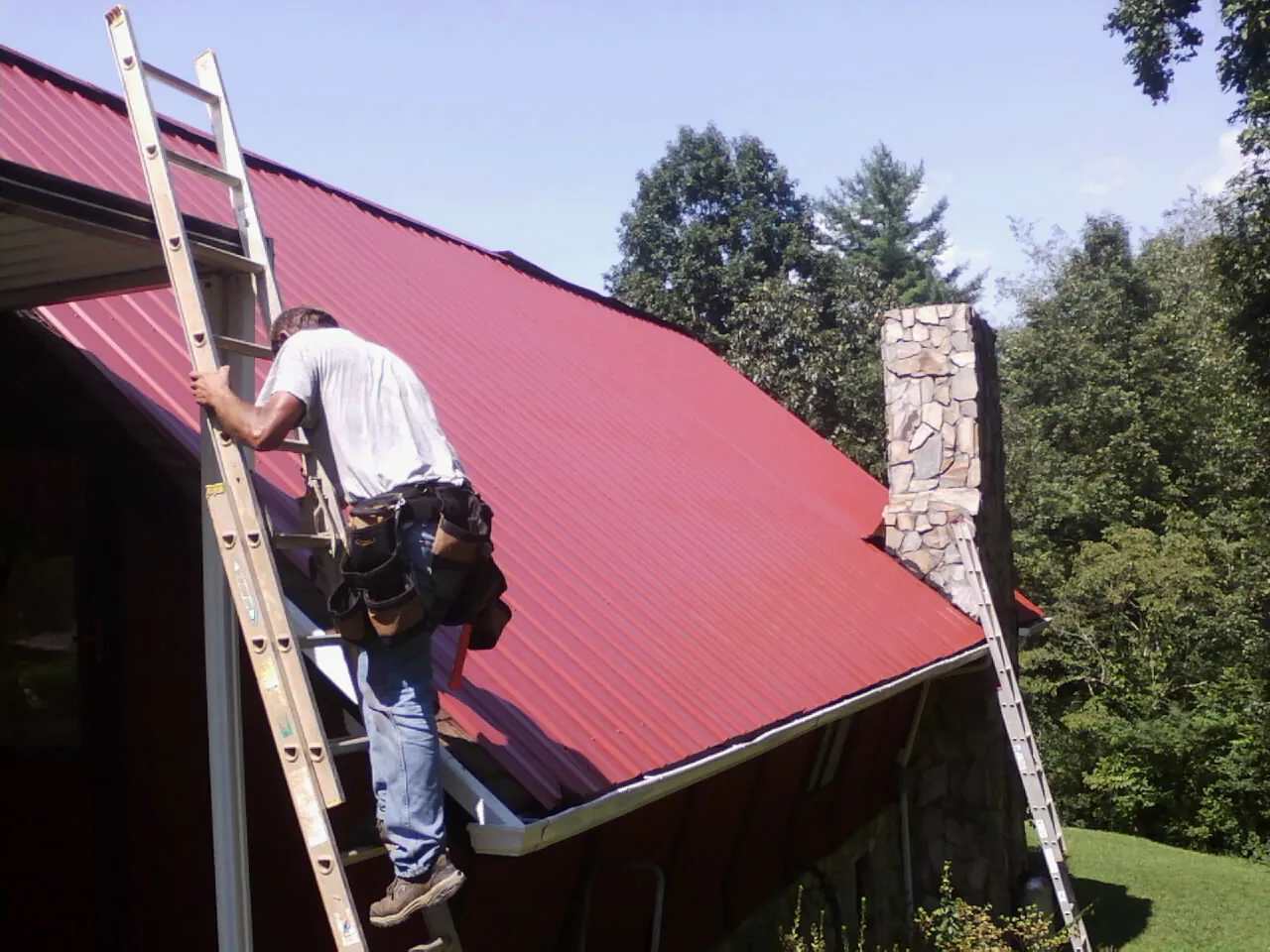 Roofer on a ladder beside a red metal roof and stone chimney — Soffit & Fascia Repair by Roofing Brothers in Reedsburg